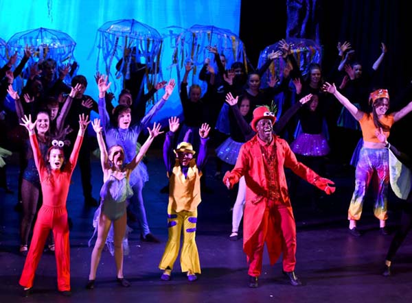 Schüler der LVS Ascot führen ein farbenfrohes Theaterstück mit fantasievollen Kostümen und Bühnenbild auf, während sie auf der Bühne tanzen und singen.